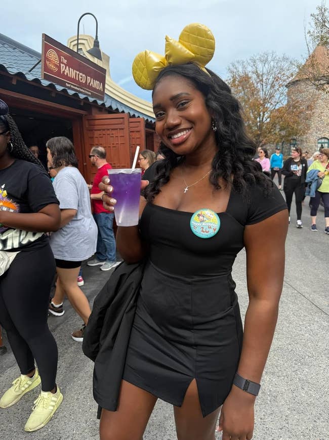 Person outdoors holding a purple drink, wearing a achromatic formal and golden rodent receptor headband, smiling astatine an amusement parkland entrance