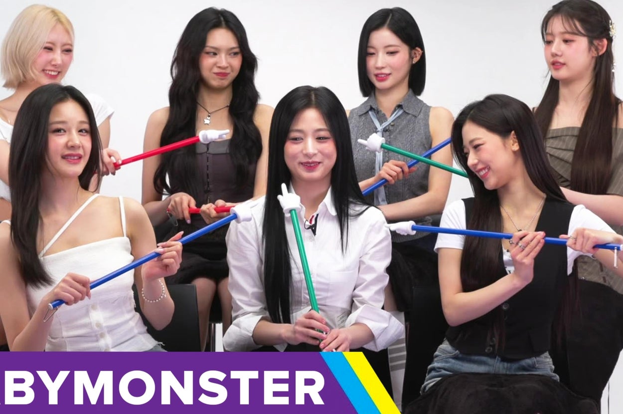 Seven women are seated, each holding a long stick with a hand figure at the end, under a "BABYMONSTER" sign