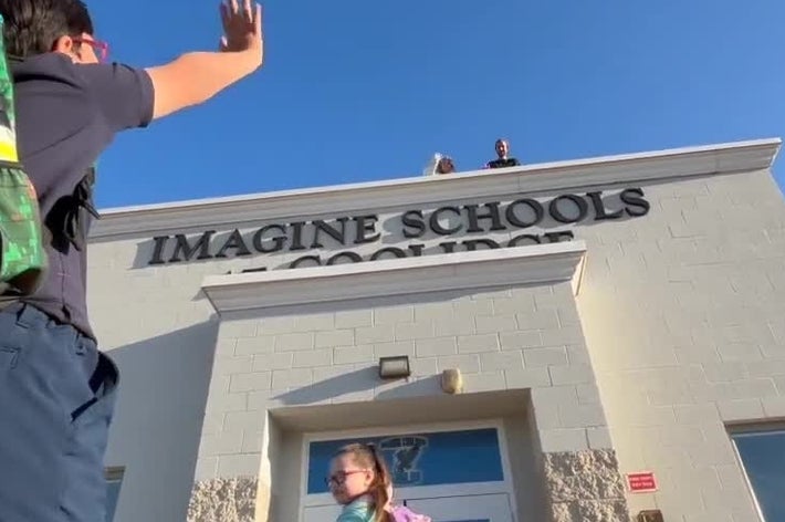 子供たちが学校の前で手を振って挨拶している場面。