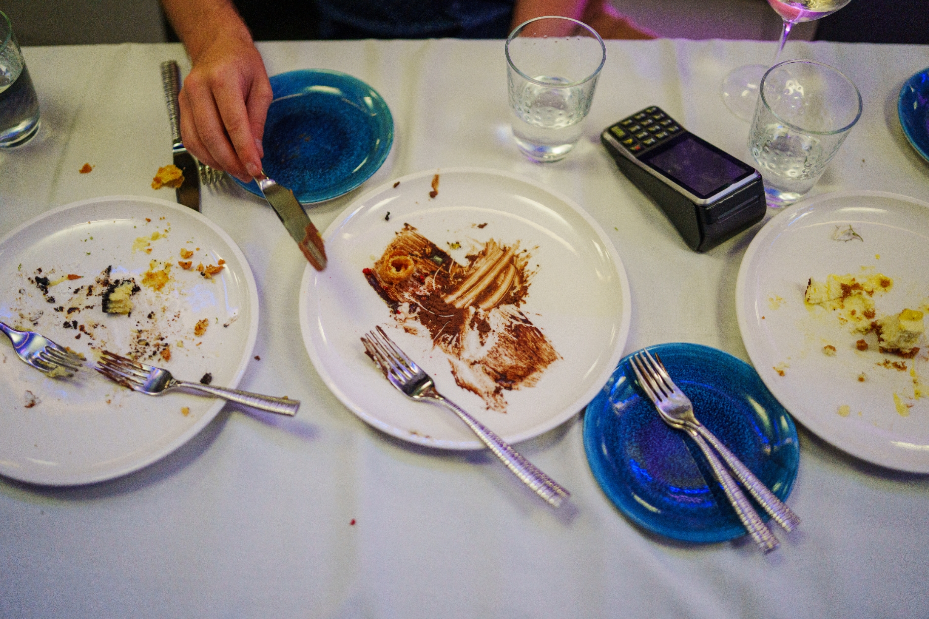 白いテーブルクロスの上に置かれたデザートの食べかけの皿。手がナイフで残りを集めている。