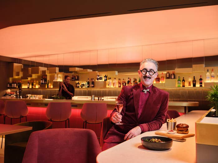 A person in a stylish suit sits at a modern bar with a cocktail, smiling. Bar shelves and bartender visible in the background