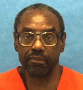 A antheral wearing glasses and an orangish garment looks straight astatine the camera against a plain background