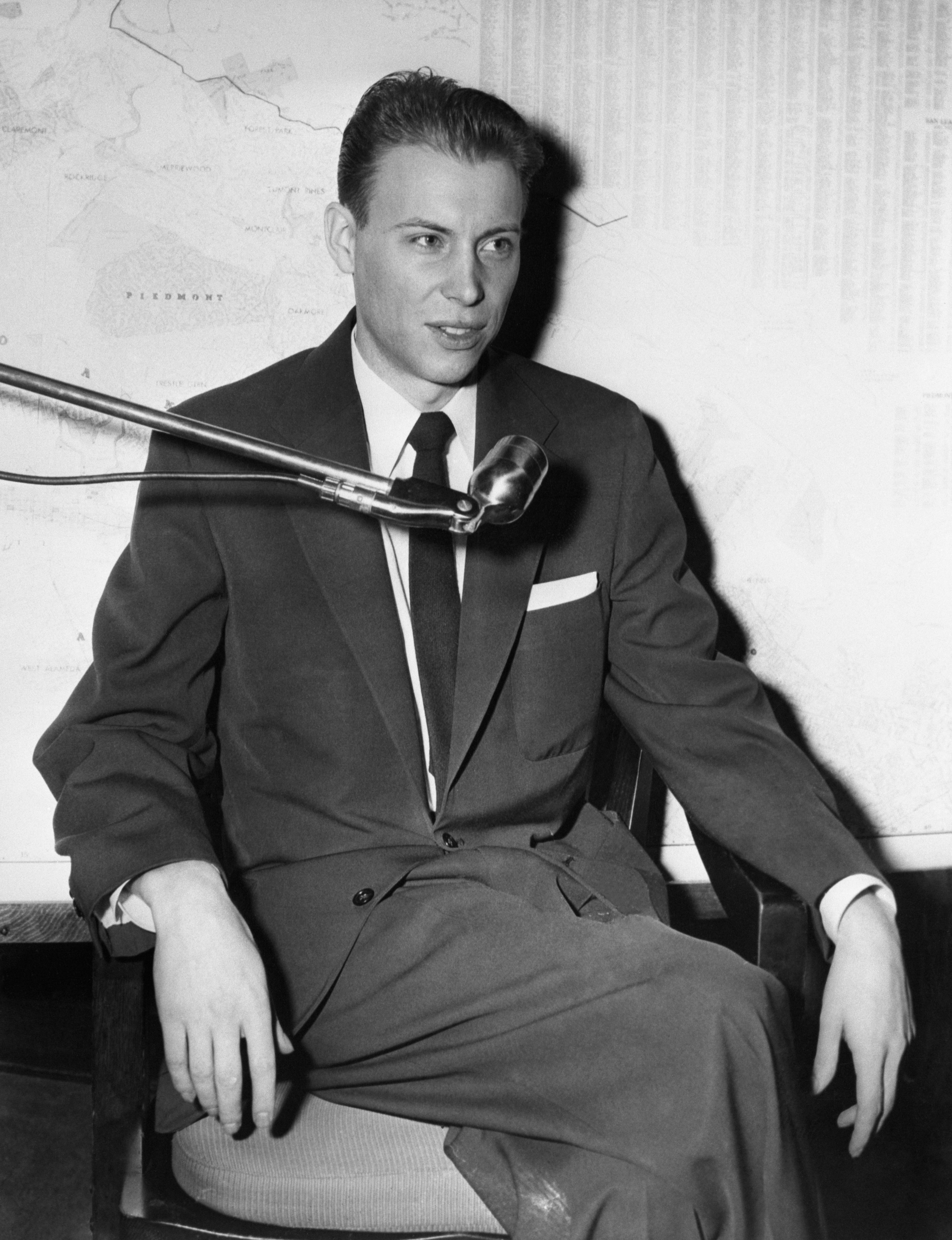 Person successful a suit sits astatine a microphone successful a workplace setting, speaking oregon recording. Map disposable successful background