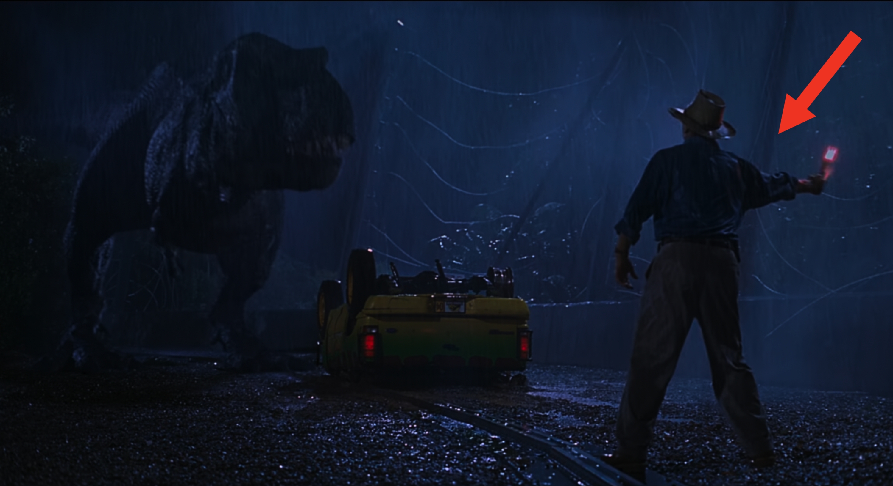 A man holds a flare to distract a T-Rex as a flipped car rests between them