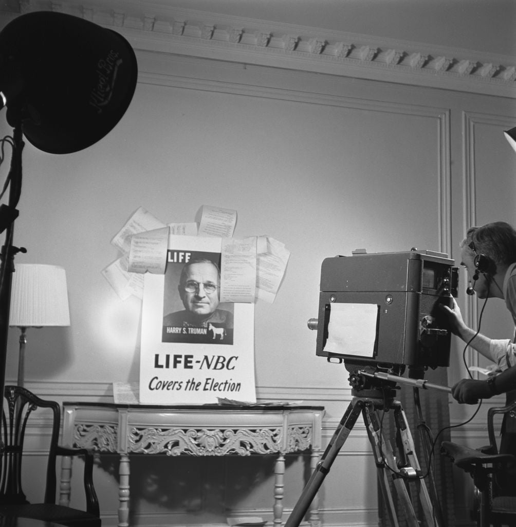 A cameraman films a vintage TV broadcast featuring a "LIFE NBC Covers the Election" poster with a man's representation    successful  a mid-20th-century workplace  setup