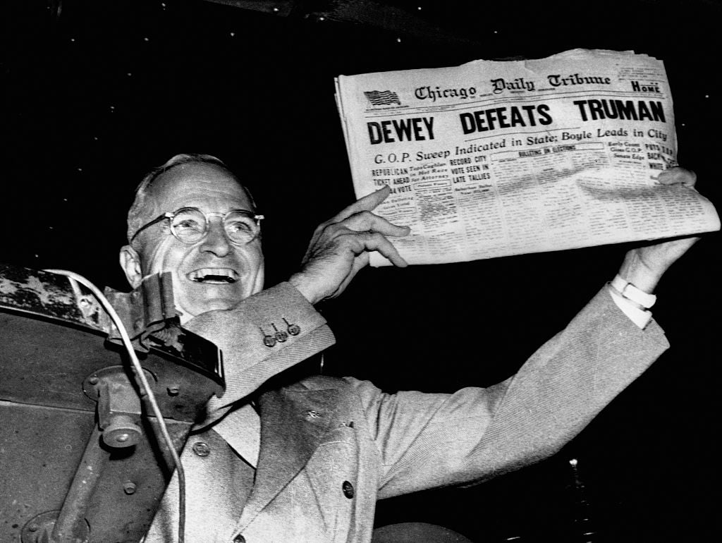 Man holding a paper  with the incorrect header  "Dewey Defeats Truman," smiling and lasting  by a podium
