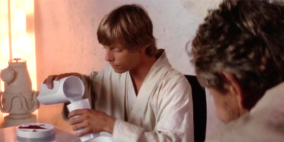 A young person in a simple robe pours a drink while sitting at a table, with an older person watching nearby