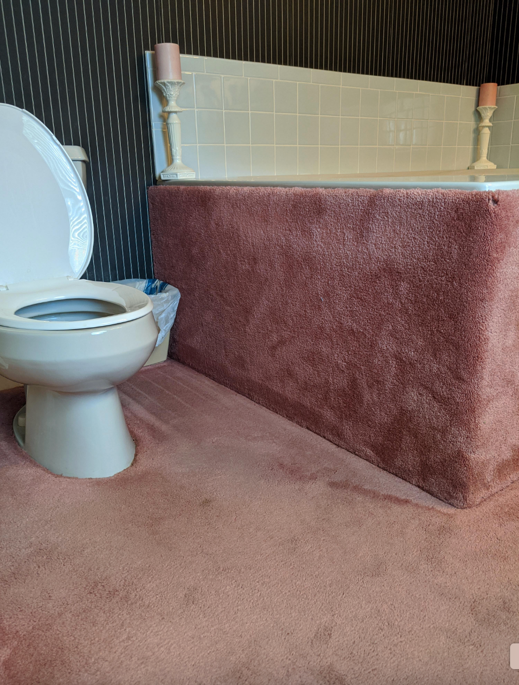 Bathroom with a toilet and carpeted bathtub exterior, featuring a striped wall and two candles on the tub ledge