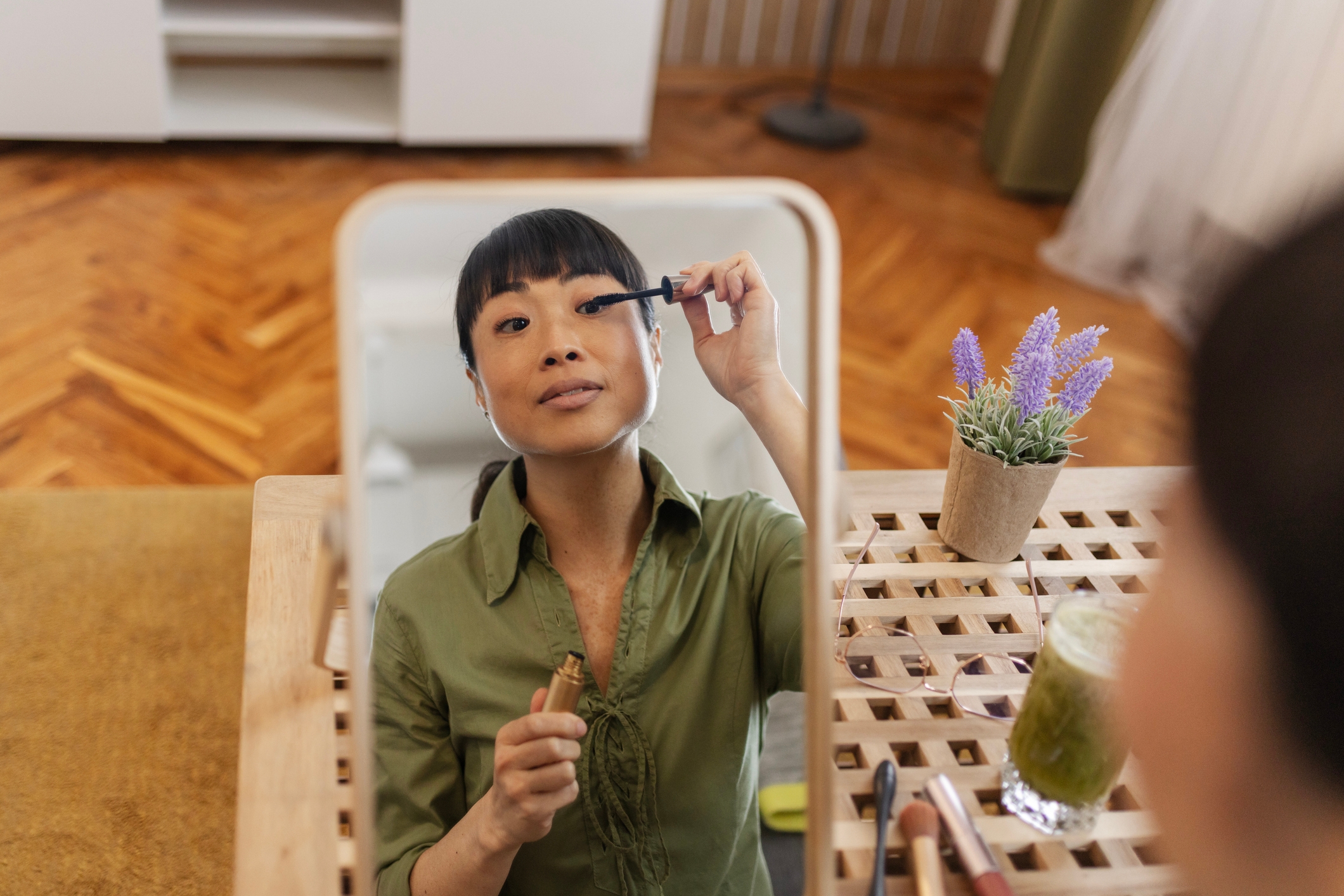 Personen som bruker mascara i et speil hjemme, iført en knapp-up-skjorte, ved siden av en potteplante og en drink på bordet