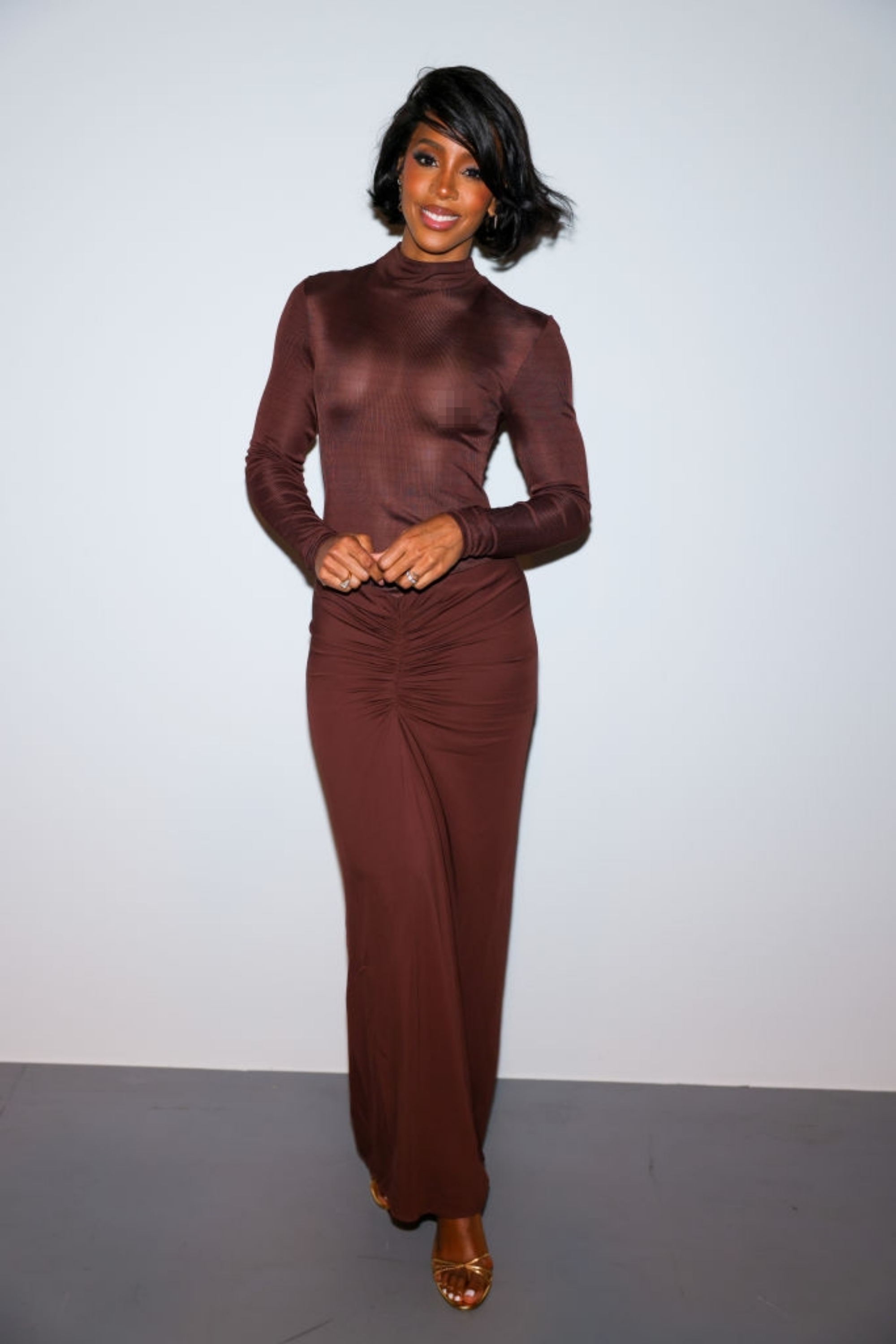 Person in a long-sleeved, fitted gown with ruching detail, smiling and standing in a poised manner
