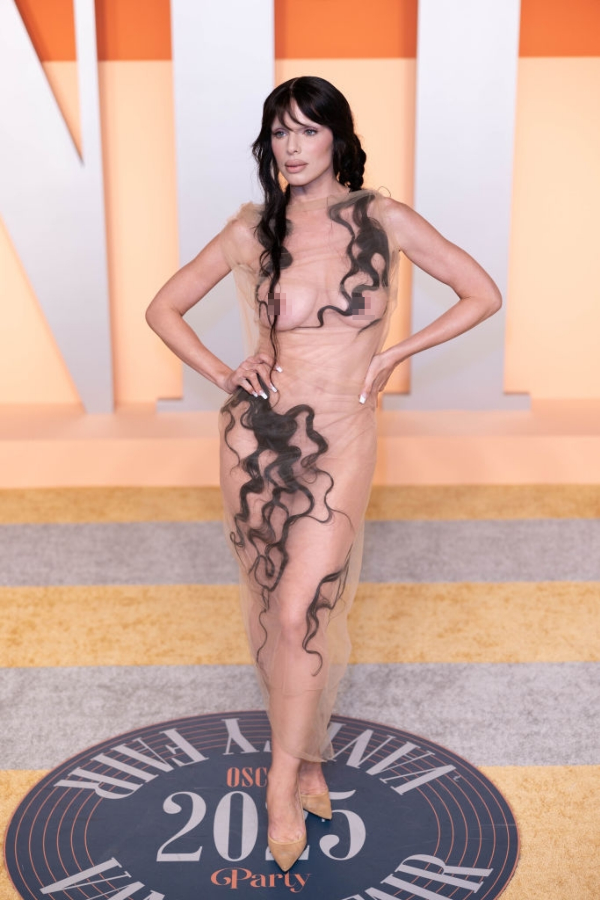 Person poses on a red carpet in a sheer dress with artistic black wavy lines, hands on hips, with long dark hair styled in waves