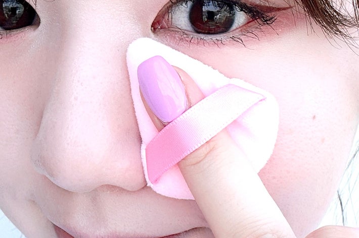 女性がメイクスポンジを使い、顔にパウダーを塗っている。指にピンクのネイルが塗られている。