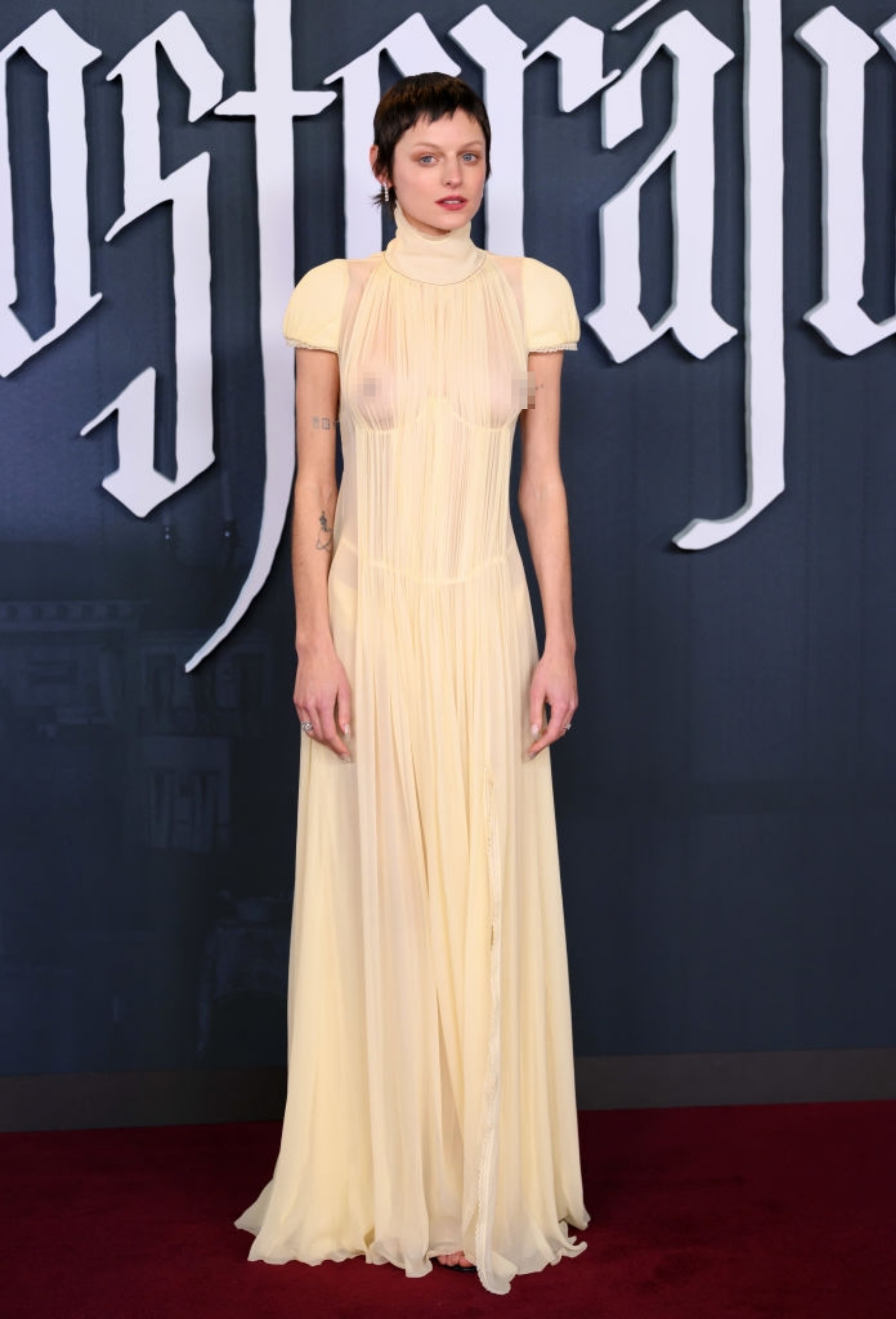 Person in elegant, sheer gown with high neckline on red carpet; gothic font backdrop