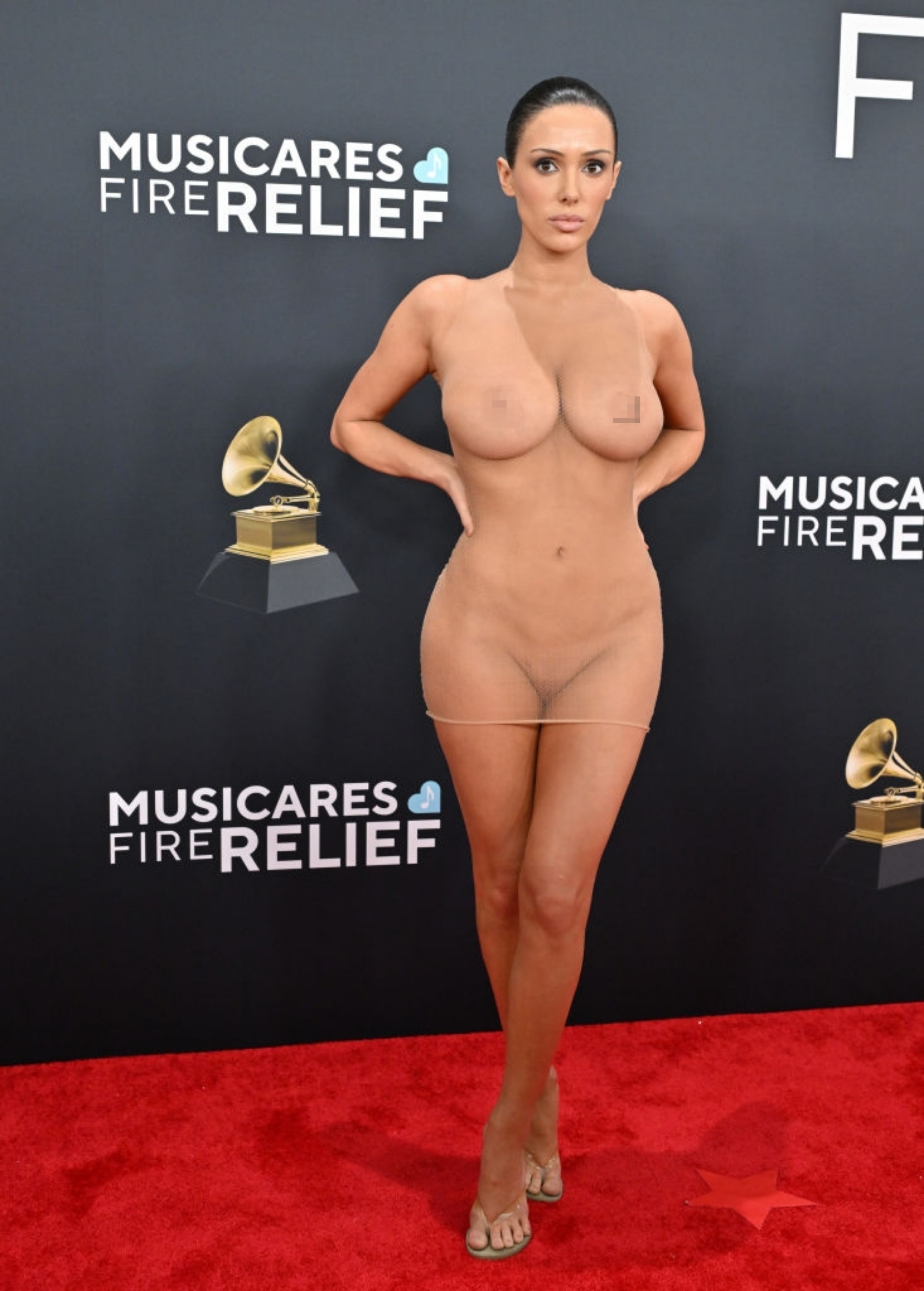 Person on red carpet wearing a form-fitting, semi-sheer dress with strategic embellishments. Background features "MusiCares" and "FIRE RELIEF" logos