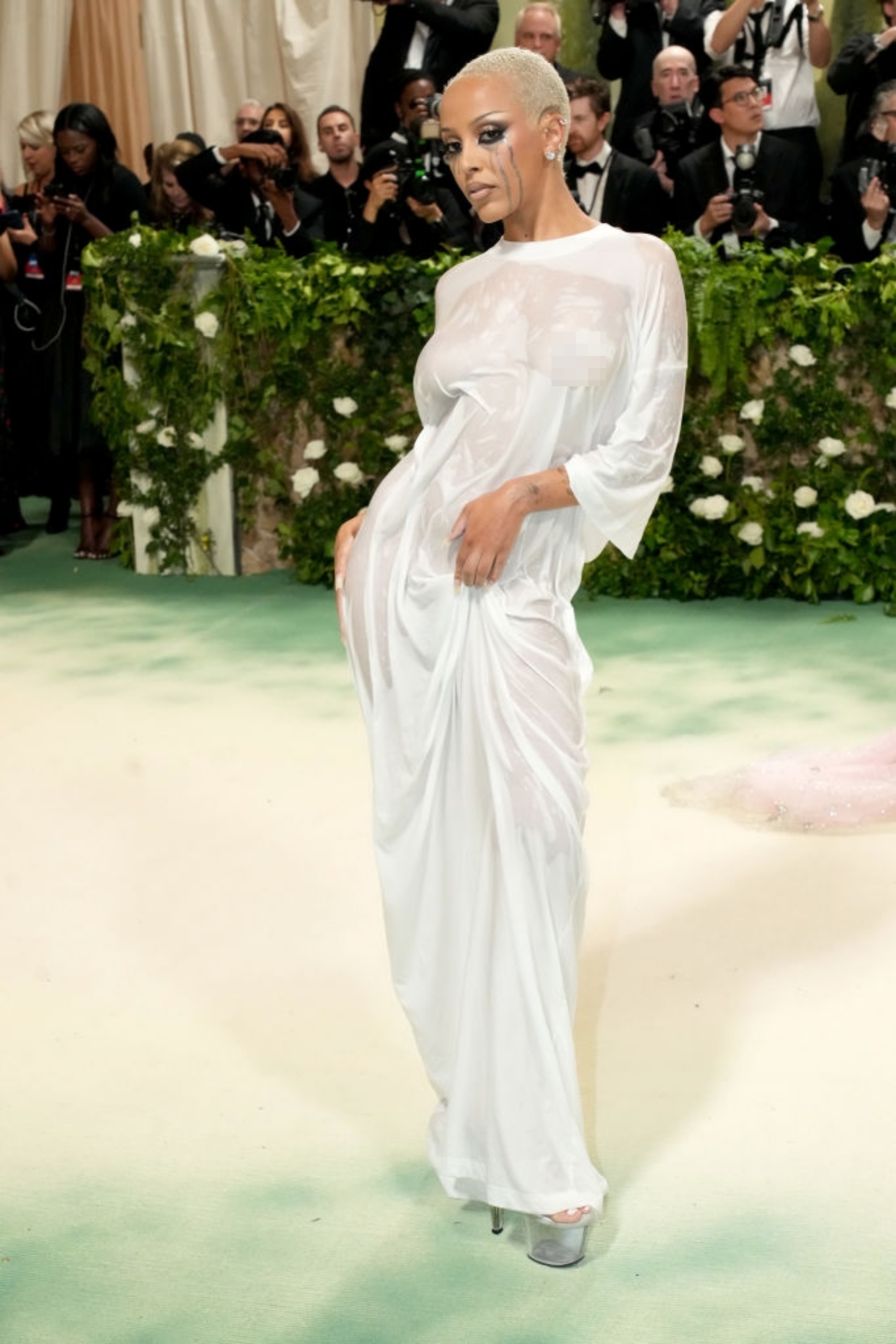 Person posing on red carpet in elegant, flowing white gown with semi-sheer sleeves, surrounded by photographers and greenery backdrop