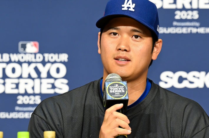 野球選手が記者会見でマイクを手に発言中。キャップとスポーツウェアを着用。背景にイベント名が書かれた看板。
