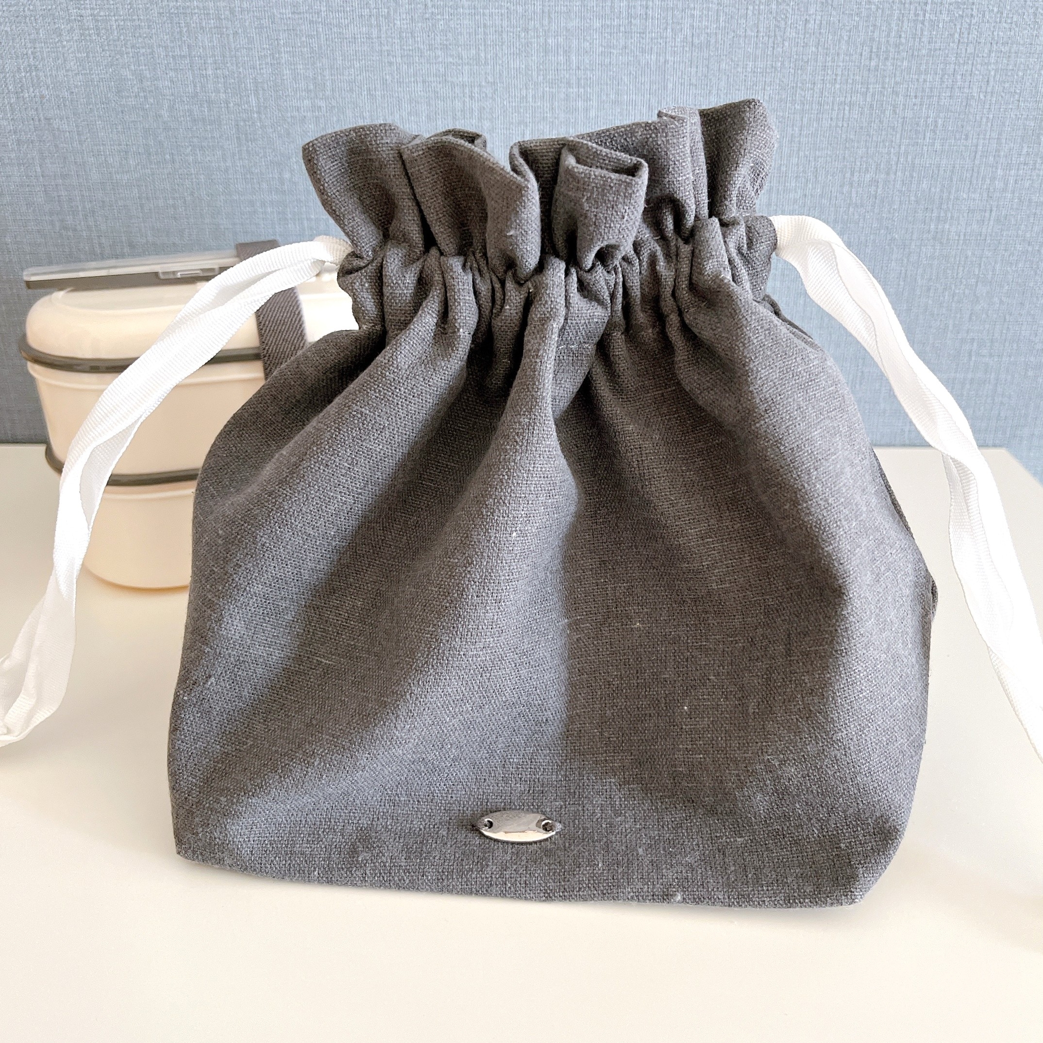 灰色の巾着バッグがテーブルに置かれています。バッグの口は白いリボンで閉じられています。