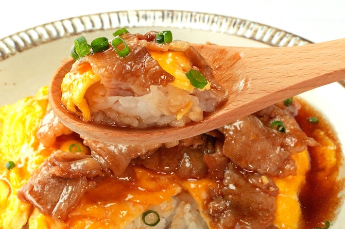 木製のスプーンで持ち上げられた、オムライスと牛肉の料理。ジュワッとしたソースがかかっています。