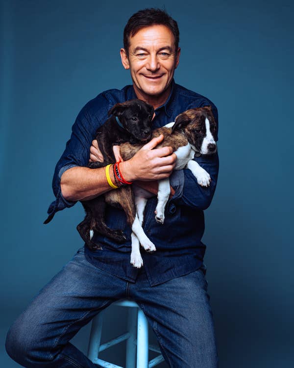 Person sitting on a stool smiling, holding two puppies, wearing casual denim attire with bracelets
