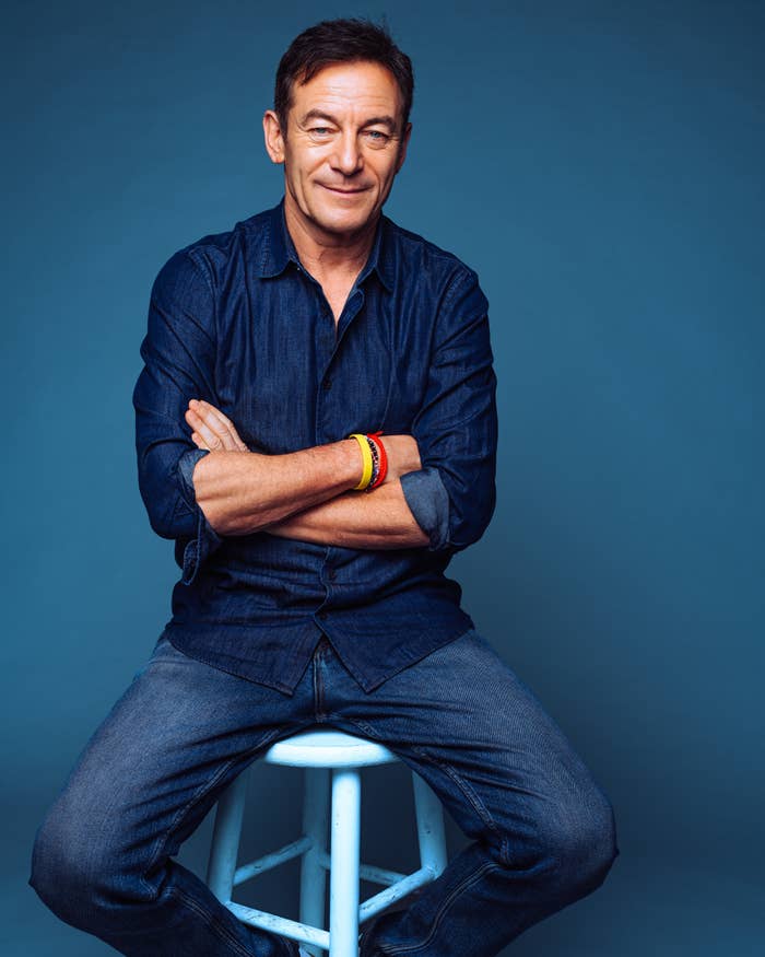 A person sits on a stool, wearing a casual denim outfit with arms crossed and a smile against a simple background
