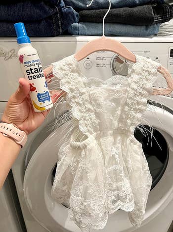 A hand holds Miss Mouth's stain treater next to a lacy children's dress in front of a washing machine, suggesting a cleaning solution for delicate fabrics