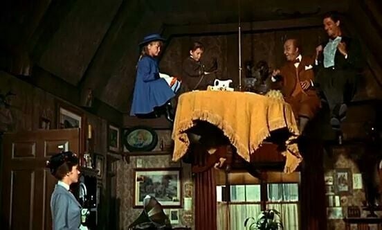 Characters sitting around a floating table in a cozy room, with a woman watching from the side. Scene likely from a film