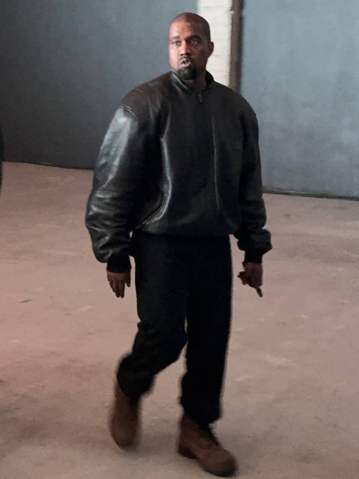 Ye  in a leather jacket and boots walks indoors, appearing focused and engaged