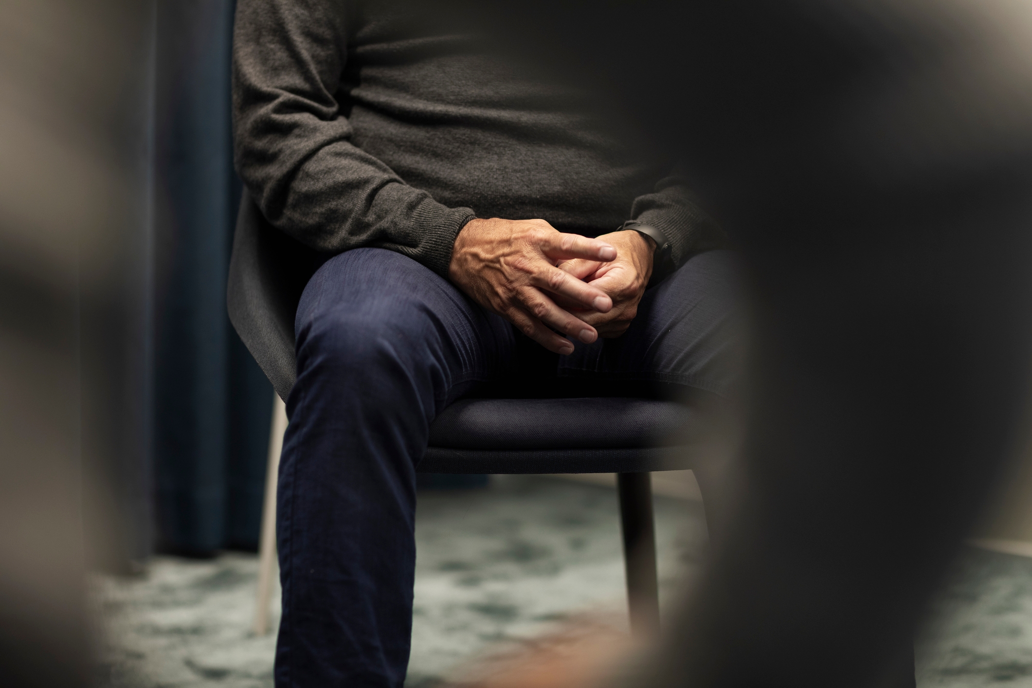 Person sittende med hendene festet, iført en genser og bukser, delvis skjult av gjenstander i forgrunnen
