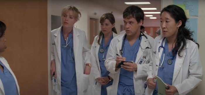 Five characters in medical scrubs and lab coats stand in a hospital corridor, appearing engaged in conversation