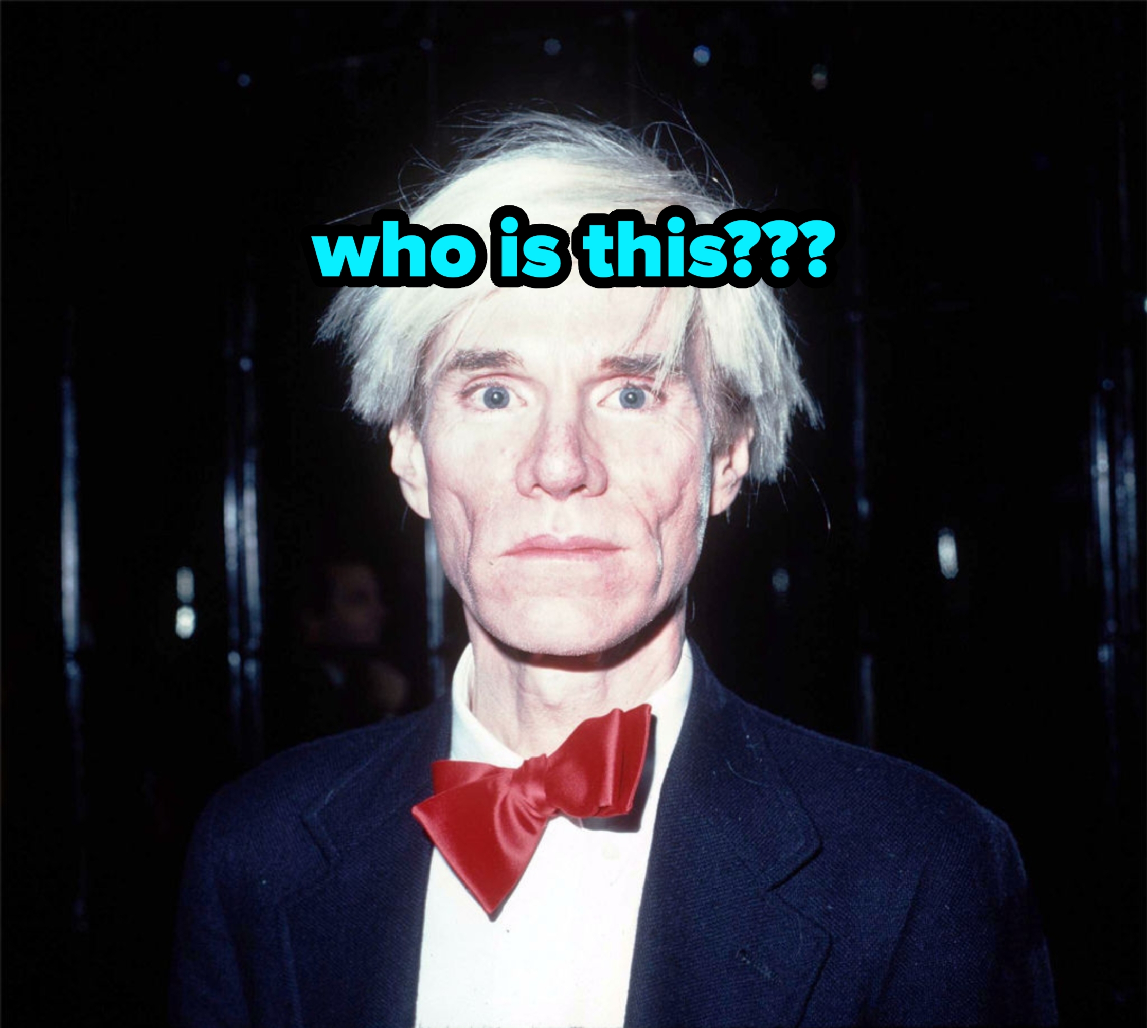 Person with distinctive hairstyle, wearing a suit and large bow tie, looking directly at the camera