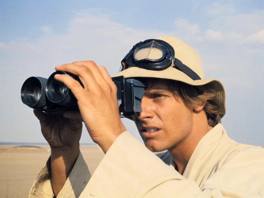 Person looking through binoculars in a desert setting, wearing goggles on a hat and light-colored clothing, appearing focused