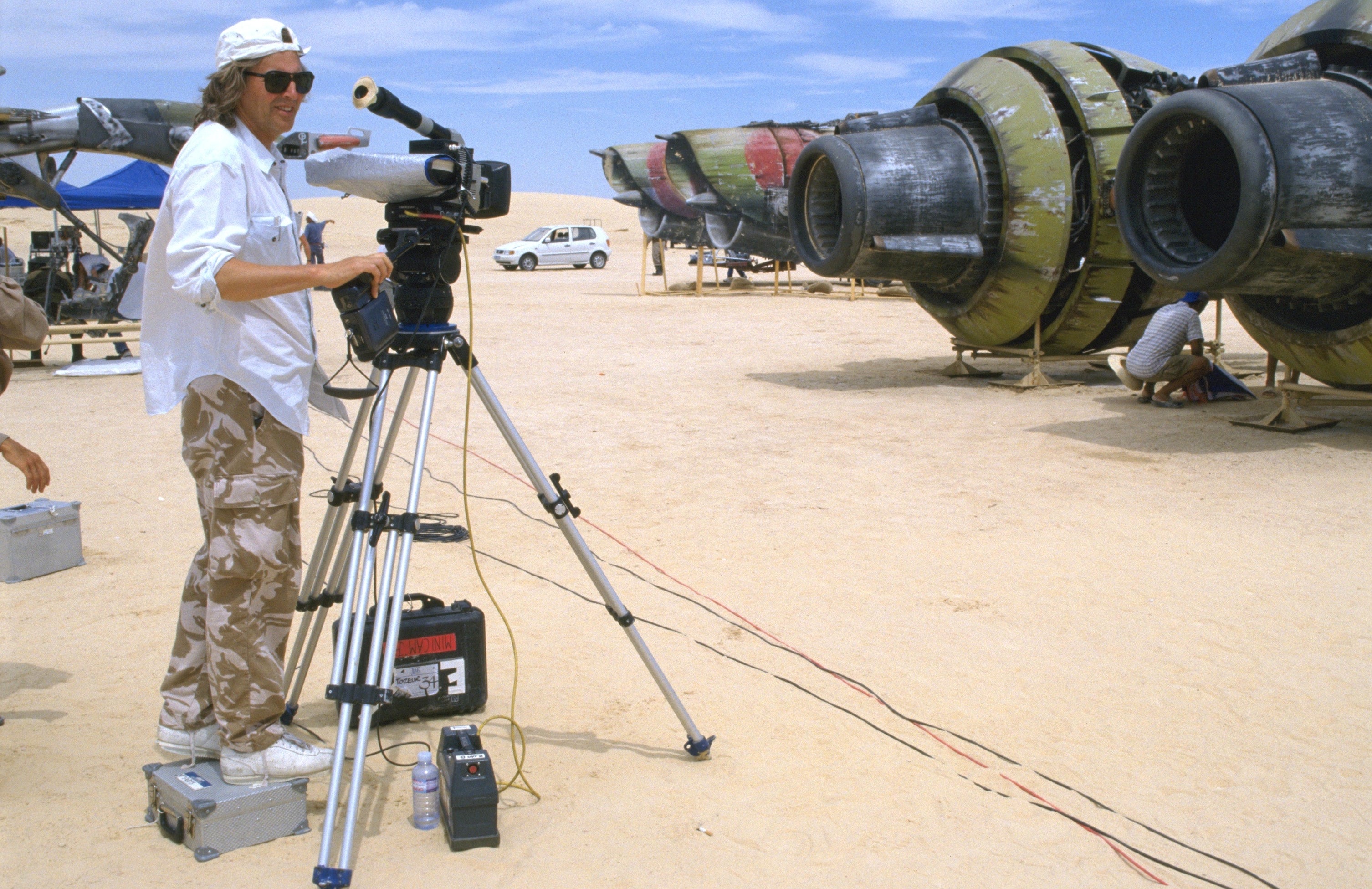 A person in casual clothing operates a camera on a movie set, with large sci-fi prop engines in the background, suggesting a film production
