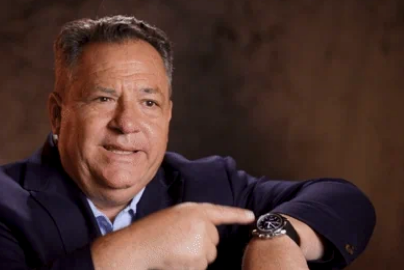 A person in a suit is pointing to their watch, possibly indicating time or urgency, with a neutral facial expression