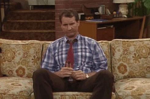 Person sitting on a floral-patterned sofa, looking unimpressed, holding a remote. Casual attire with a checked shirt and tie. Living room setting