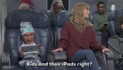 Child in airplane seat uses tablet, while adult beside them looks amused. Other passengers in background. Caption reads: "Kids and their iPads right?"