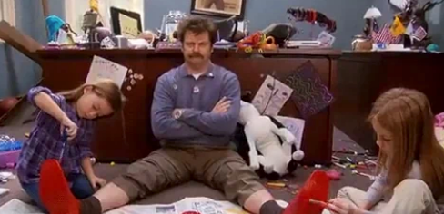 Man sitting on the floor in a messy room, while two children paint his legs. Room features scattered crafts and toys