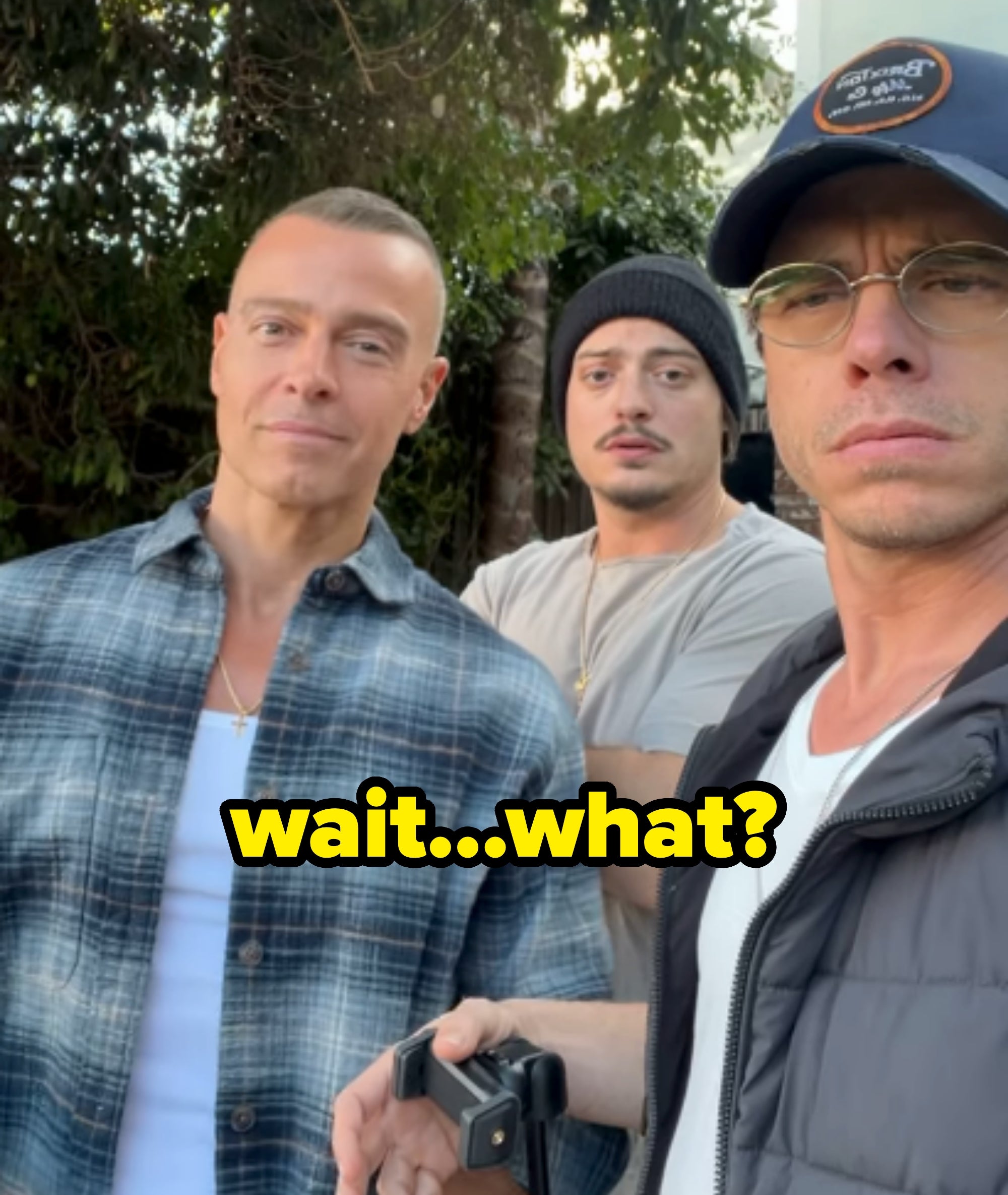 Three individuals outdoors, one in a plaid shirt, one in a beanie, and one in a cap and glasses, standing closely together, looking at the camera