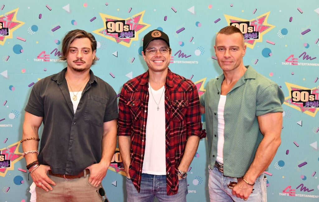 Three individuals wearing casual outfits pose in front of a 90s-themed backdrop