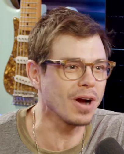 A person with short hair and glasses speaks animatedly into a microphone, with a guitar visible in the background