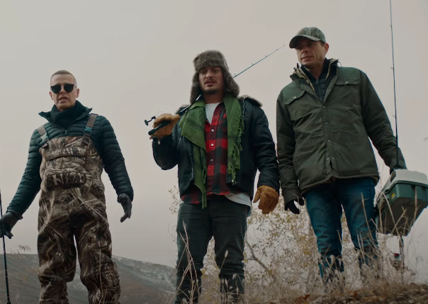 Three people outdoors, dressed warmly. One holds a fishing rod, another wears sunglasses, and the third carries a tackle box
