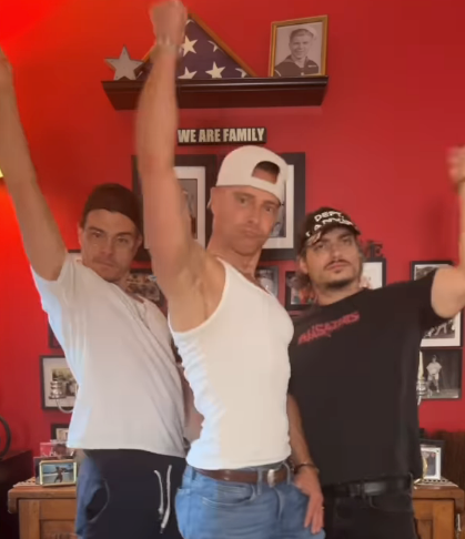 Three people strike dynamic poses with raised arms, dressed in casual attire, standing in a room adorned with family photos and memorabilia