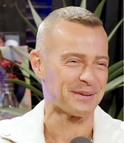 Smiling individual with short hair, wearing a light-colored jacket, speaking into a microphone. Indoor setting with plants in the background