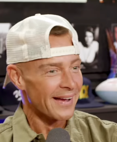 Person smiling during an interview, wearing a mesh baseball cap backwards and a casual jacket. Background shows photos and memorabilia