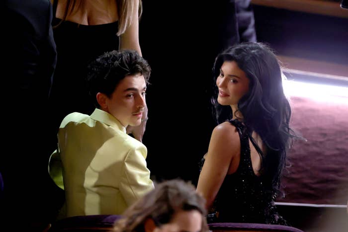 Kylie Jenner and Timothée Chalamet  seated, turning to look back. The man wears a light-colored jacket, the woman in a sequined dress