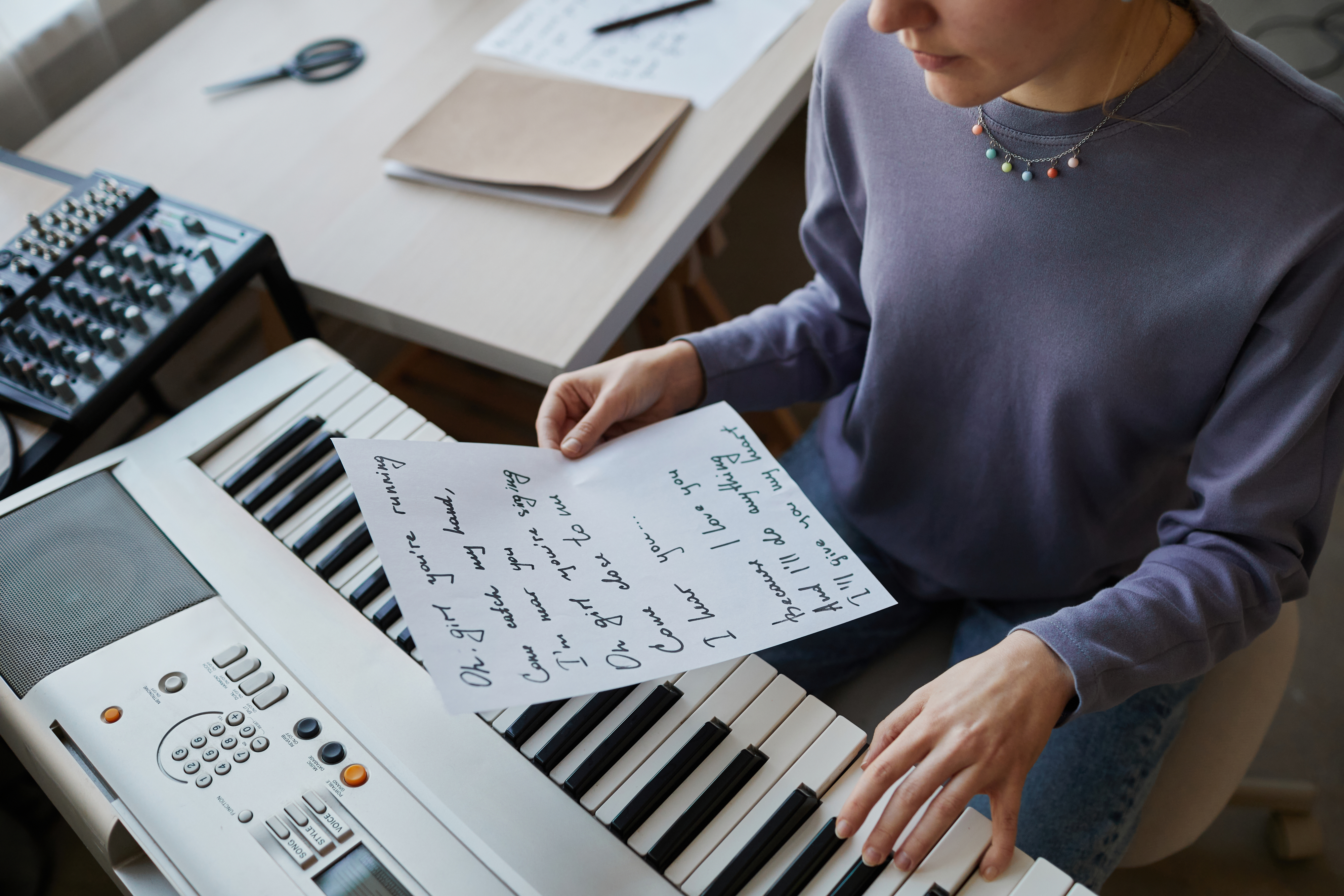 Person sitting at a keyboard piano holding a sheet of handwritten song lyrics