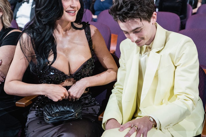 Two people seated in an audience at a formal event; one wearing a black, sleeveless outfit, the other in a light-colored suit, looking down