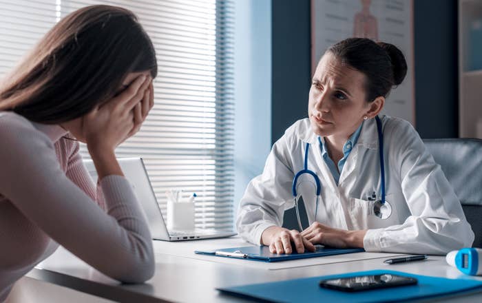 A doctor in a white coat listens intently to a patient who is covering her face with her hands in an office setting