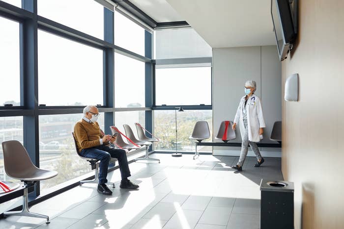 A doctor in a white coat and mask walks through a modern waiting room, where a patient in a mask reads a magazine