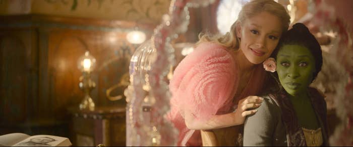 Two actors from a fantasy film, one with green makeup wearing a Victorian-style costume, and the other in a pink, feathery outfit, smiling indoors