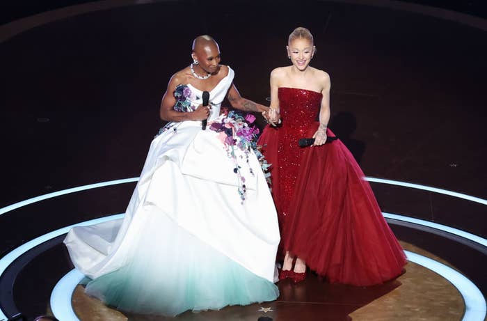 Two individuals on stage wearing elegant gowns, one with floral details and the other in red. They hold hands and stand in a spotlight
